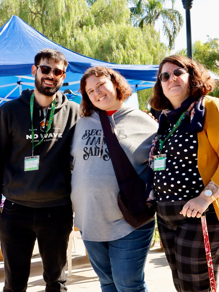 Tres personas posan al aire libre, con árboles y un toldo azul detrás.